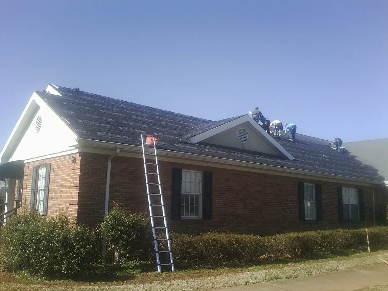 Crew tearing shingles off a brick ranch roof with a ladder set — Metal Roof Repair work by our crew in Hartland